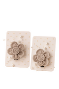 Pair of beige flower-shaped earrings on white cards with gold polka dots, placed on a beige surface with a plant in the background.